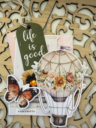 Decorative card with 'life is good' tag, butterfly, and floral design on a decorative wooden background.