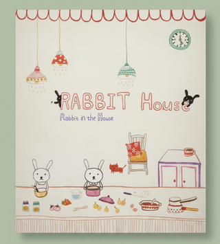 A children's book titled 'Rabbit House: Rabbit in the House' with a whimsical hand-drawn illustration on the cover, photographed flat against a dark brown wooden background.