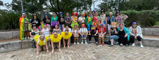 A large group of people posing together outdoors on a paved stone patio, dressed in colorful carnival or clown costumes with bright yellow ruffles, hats, and masks. Trees and shrubs are visible in the background.