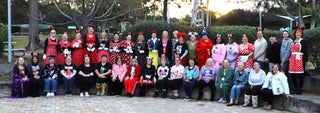 A large group of people posing together outdoors in front of trees and a park setting, wearing various colorful costumes and themed outfits.